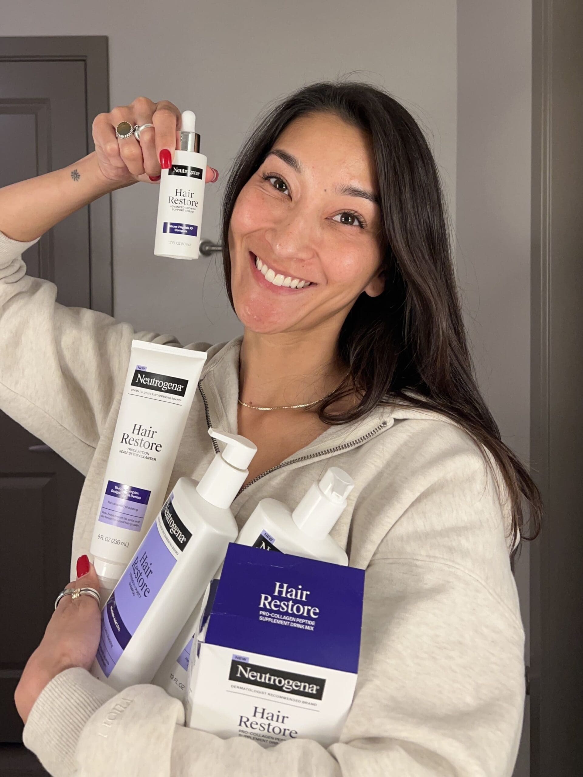 Taylor Nakakihara holding the Neutrogena Hair Restore line products, including the shampoo, conditioner, scalp cleanser, serum, and collagen supplement, while smiling in a well-lit bathroom.
